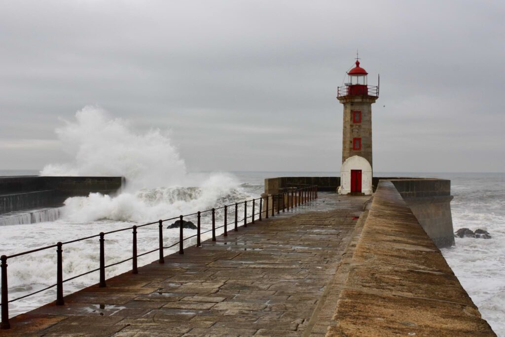 Leuchtturm am Ende eines Steinwegs mit links einem Geländer und hoher Brandung links im Bild.